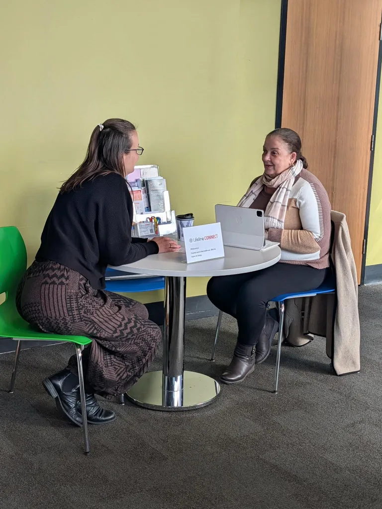 Two people talking at a small round table at the Connect Hub, beside a Lifeline Connect welcome card and a rack of support brochures.